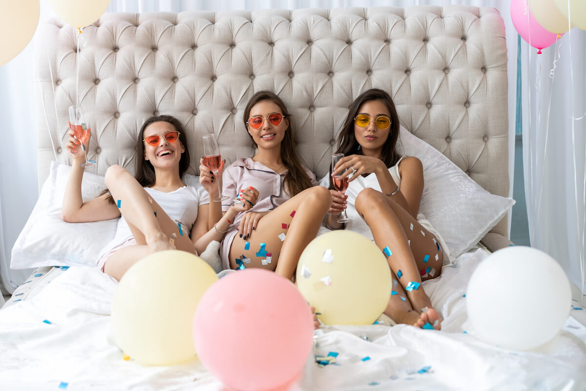 three women sitting in a bed with glasses of wine at a slumber party