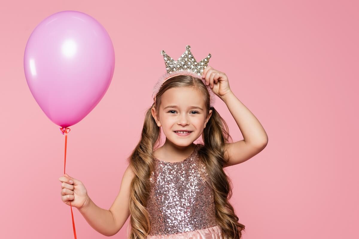 pretty little girl in fancy dress holding a pink balloon and putting on a crown headband.