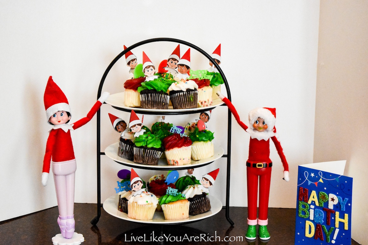 two elf on the shelf scout elves standing next to a cupcake stand with decorated red and green cupcakes.