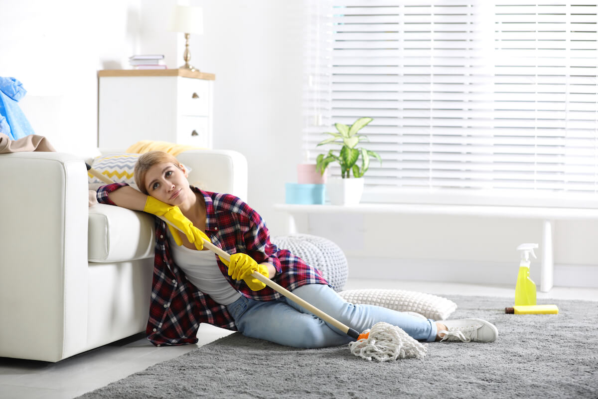 woman feeling overwhelmed by cleaning her house, sitting on lounge room floor with mop