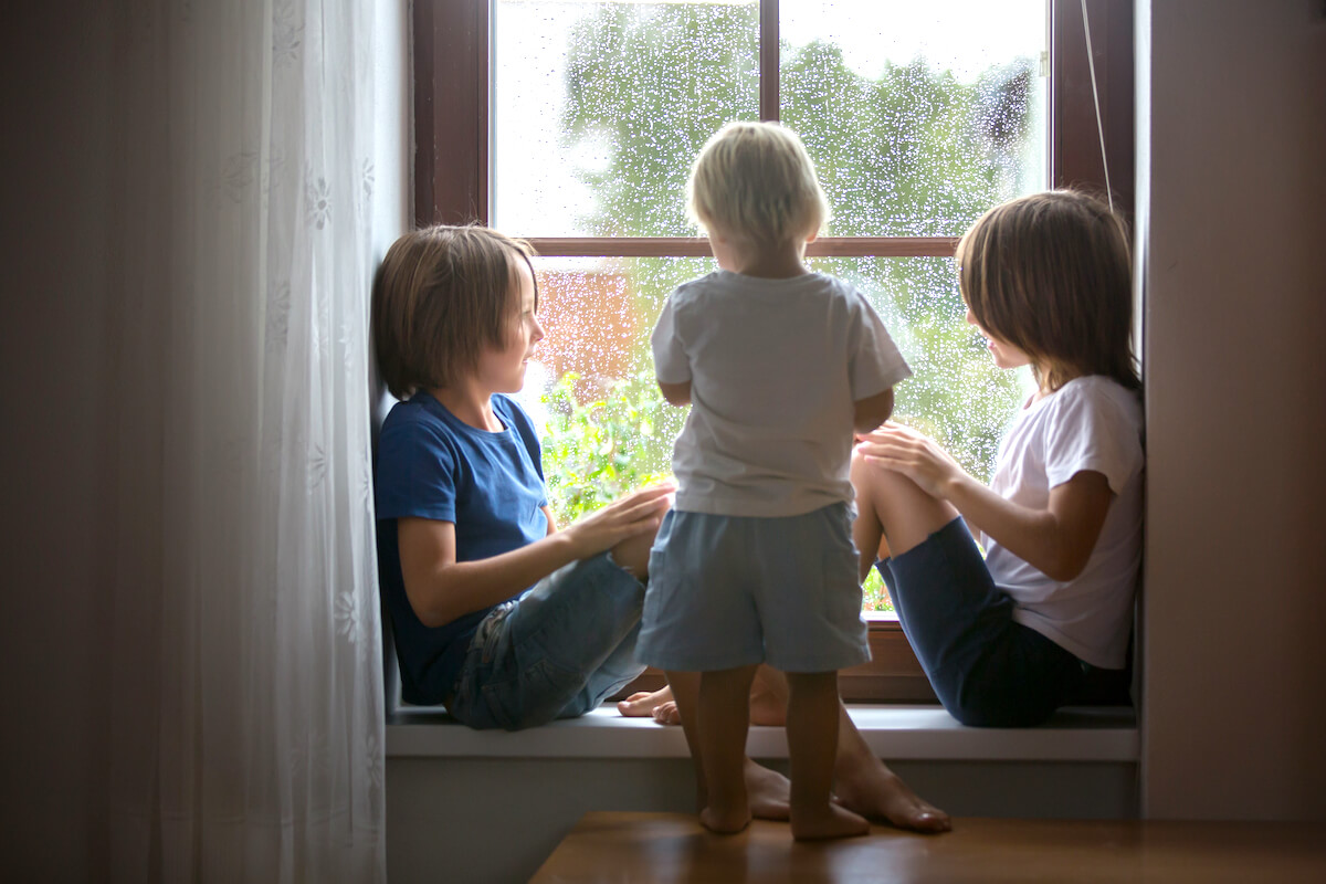 three young boys sitting on the window seat inside their house looking out at the rain