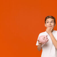 Teen boy with piggy bank dreaming about some things he can buy, orange studio background with empty space