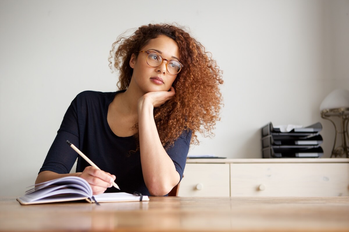 curly haired woman sitting at a table indoors reflecting and writing in a journal.