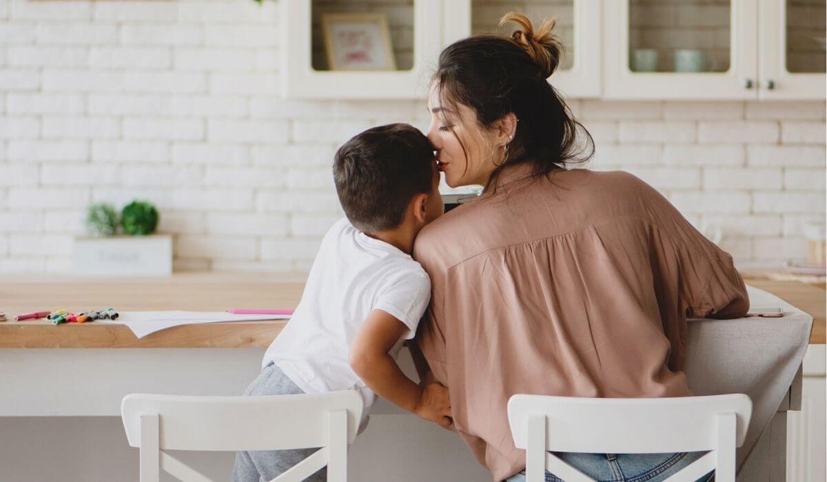 woman kissing son on forehead as they sit at kitchen bench