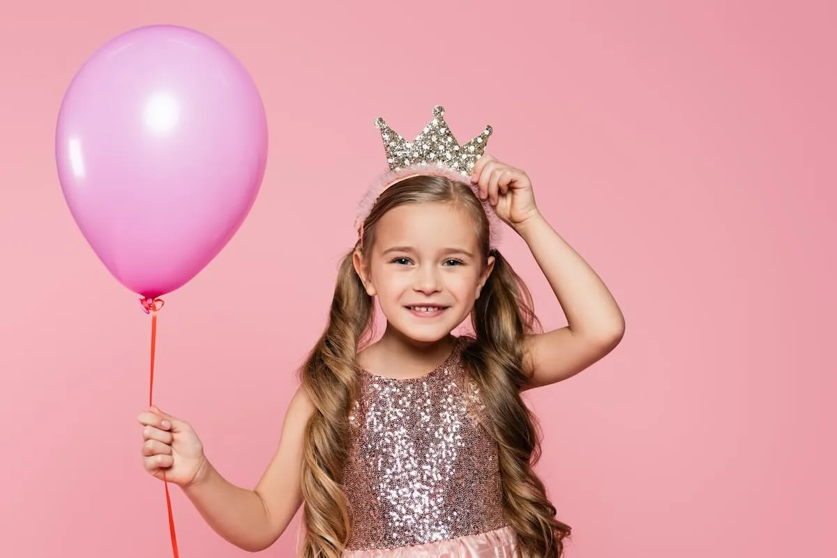 pretty little girl in fancy dress holding a pink balloon and putting on a crown headband.