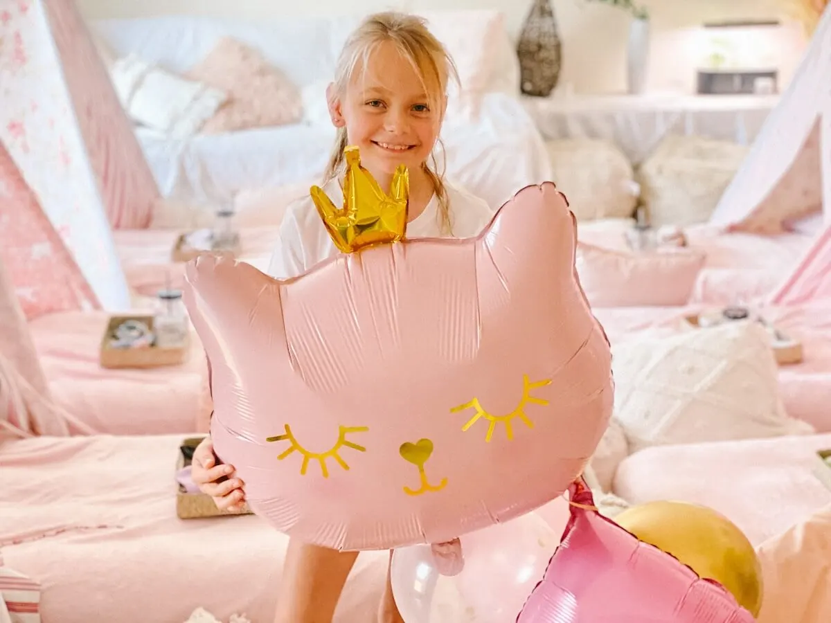 excited little girl holding a large pink cat balloon in front of a teepee birthday party set up.