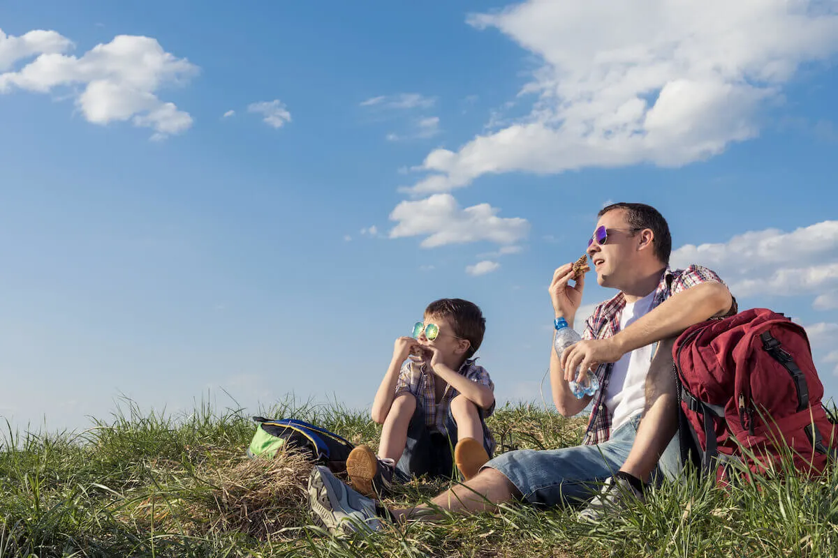 father and son sitting in the grass eating lunch while on a day hike