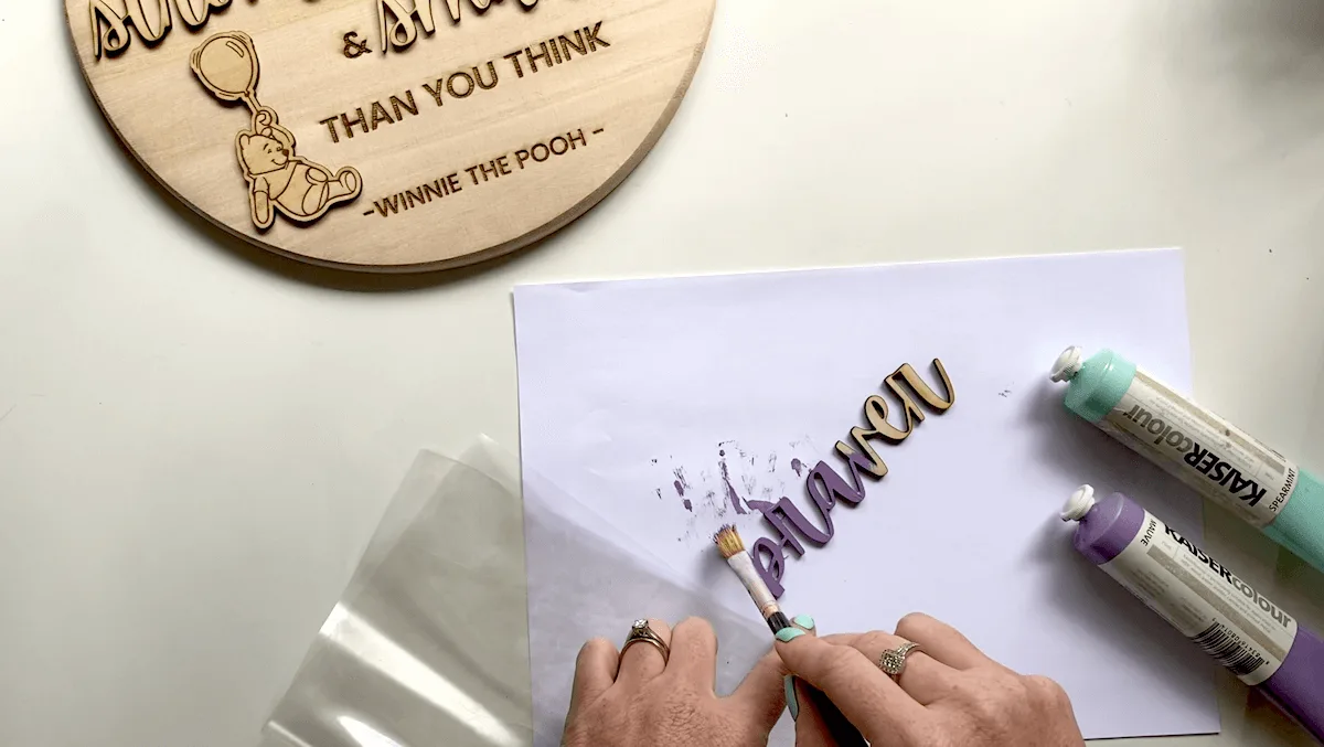 person painting wooden words on a white desk with purple paint and a wooden sign in background