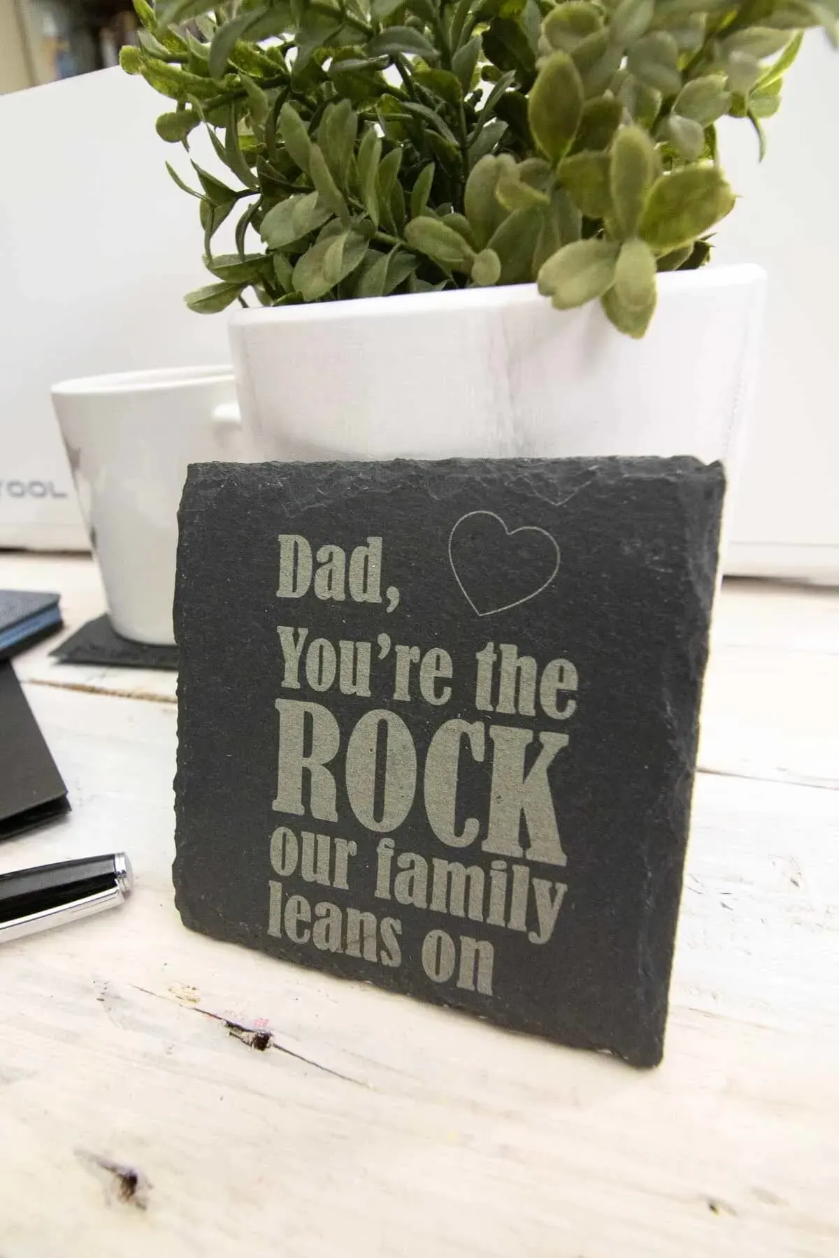 slate coasters engraved with dad you're the rock our family leans on resting against a pot plant